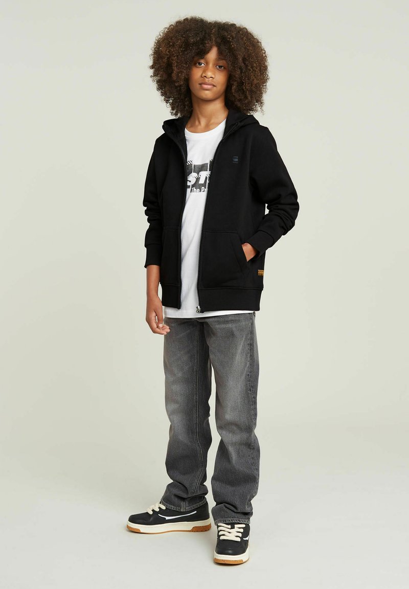 Sudadera negra con cremallera y bolsillos sobre una camiseta gráfica blanca, combinada con jeans grises y zapatillas negras con suela crema.