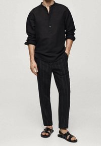 Maglietta henley nera a maniche lunghe con scollatura a bottoni, abbinata a pantaloni neri a righe sottili e sandali neri. Stile semplice e monocromatico.