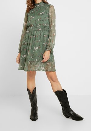 Kobieta w zielonej, kwiatowej sukience z długim rękawem i falbanami u dołu, nosząca czarne kowbojki do połowy łydki, stojąca z jedną nogą lekko wysuniętą do przodu.