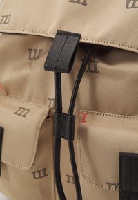 Zaino in tela beige con hardware nero, numerose tasche e dettagli del logo stampati in grigio scuro e rosso. Dettaglio in tessuto testurizzato visibile.