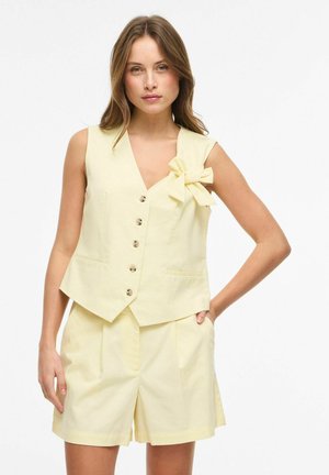 Kvinne iført blekgul ermeløs knappet vest med sløyfedetalj og matchende høytlivsshorts, poserer med hånd i lommen.