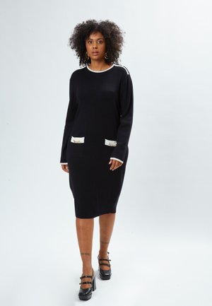 Robe tricotée noire à manches longues, avec des accents blancs au niveau du col et des poches à motifs. Portée avec des chaussures noires à talons carrés.