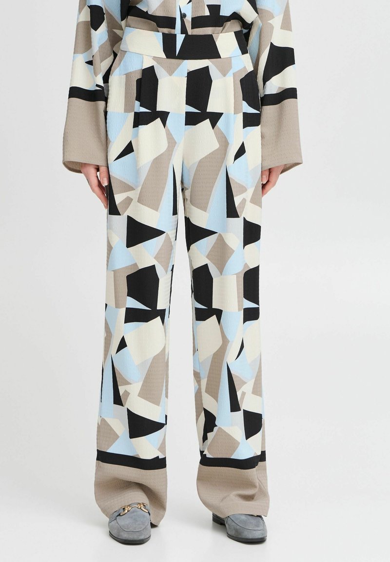 Pantalon taille haute, coupe large, avec des motifs géométriques abstraits beige, noir, bleu clair et blanc, porté avec des mocassins gris.