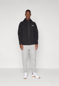 Haut à capuche zippé noir au design structuré, avec un logo sur la poitrine gauche. Associé à un pantalon de survêtement gris clair et des baskets blanches.