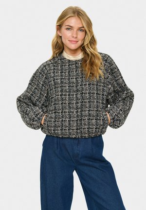 Femme blonde avec des vagues lâches, portant un pull texturé noir et beige et un pantalon bleu taille haute, les mains dans les poches, se tenant devant un fond blanc.