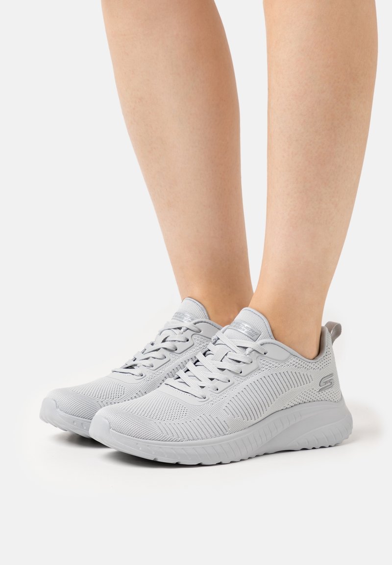 Pieds portant des baskets tricotées gris clair avec des lacets et des semelles blanches épaisses, présentés sur un fond blanc uni.