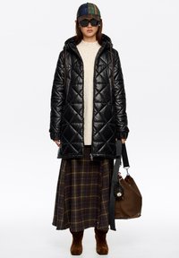 Czarna pikowana kurtka puchowa z kapturem, beżowy sweter z dzianiny, spódnica w kratę, brązowa torba oraz brązowe zamszowe botki; dodatkowo czapka w kratę i okulary przeciwsłoneczne.