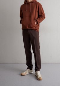 Sweat à capuche marron avec poche kangourou, associé à un pantalon marron foncé effilé. Les chaussures combinent des textures beige et blanche.