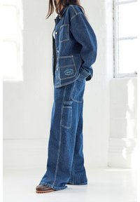 Jeansoutfit med en knäppbar jacka och vida byxor, båda i mörkblått med vita smala ränder och synliga sömmar.