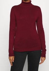 Burgundyröd polotröja i lätt stickning, figursydd design, långa ärmar och ribbad nederkant och ärmslut; visas ihop med svarta byxor.