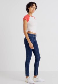 Bílý a červený zkrácený tričko v kombinaci s tmavě modrými Skinny džínami. Model má na sobě bílé tenisky. Jednoduchý design, krátké rukávy a přiléhavý střih.