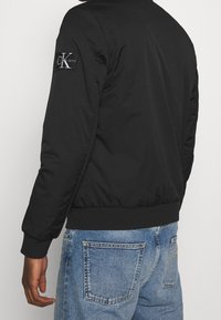 Giacca bomber nera con polsini e orlo a costine, presenta una patch con logo sulla manica sinistra e un materiale liscio e strutturato. Indossata con jeans di denim.