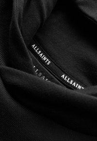 Tela negra con un acabado texturizado y acanalado. Presenta bordes suaves y etiquetas blancas de "ALLSAINTS" visibles en los pliegues.