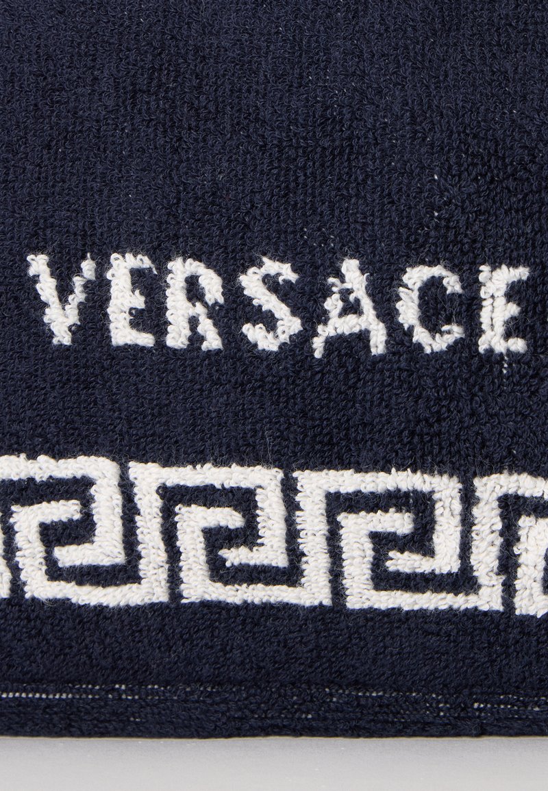 Tissu texturé bleu foncé avec texte blanc "VERSACE" et un motif clé grecque blanc sous le texte.
