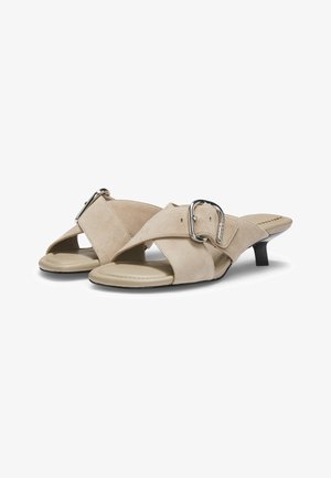 Sandalias mule de ante beige con punta abierta, tiras cruzadas y detalles de hebilla plateada, con tacones bajos tipo gatito.
