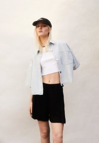 Lichtblauwe spijkerjack, korte mouwen, knoopsluiting, gedragen over een witte crop top; gecombineerd met zwarte shorts van gladde stof. Zwarte pet op.