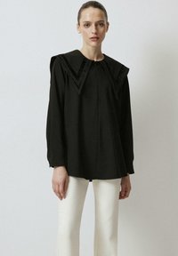 Top nero con un ampio colletto a V, caratterizzato da dettagli testurizzati. Maniche lunghe e silhouette a forma di A, abbinato a pantaloni bianchi.