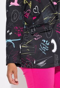 Blazer noir avec du texte et des motifs de style graffiti multicolores. Détail d'un revers et fermeture à un seul bouton, associé à un pantalon rose.