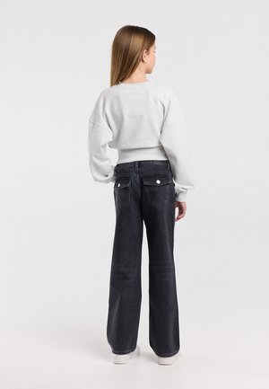 Jong meisje van achteren gezien, draagt een lichtgrijze cropped trui, zwarte wijde jeans met achtervleugeltassen en witte sneakers.