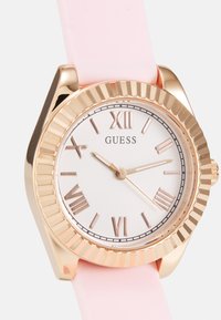 Montre dorée avec un cadran blanc, un bracelet en silicone rose et des chiffres romains. La lunette texturée ajoute un intérêt visuel. Design simple et fonctionnel.