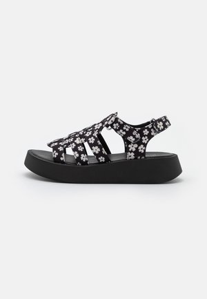 Sandales noires à lanières avec un motif floral blanc, semelle noire épaisse et fermeture à boucle réglable pour un maintien sécurisé.