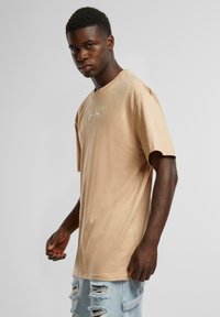 Beige cotton t-shirt with short sleeves, relaxed fit, and a tonal logo printed on the front; worn with distressed light blue jeans.