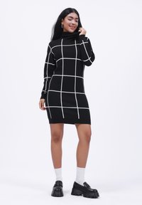 Melnas turtleneck kleitas ar baltu režģa rakstu, piegulošs dizains, ribota apkakle un izliektas maliņas. Modelis valkā masīvas melnas kurpes un baltas zeķes.