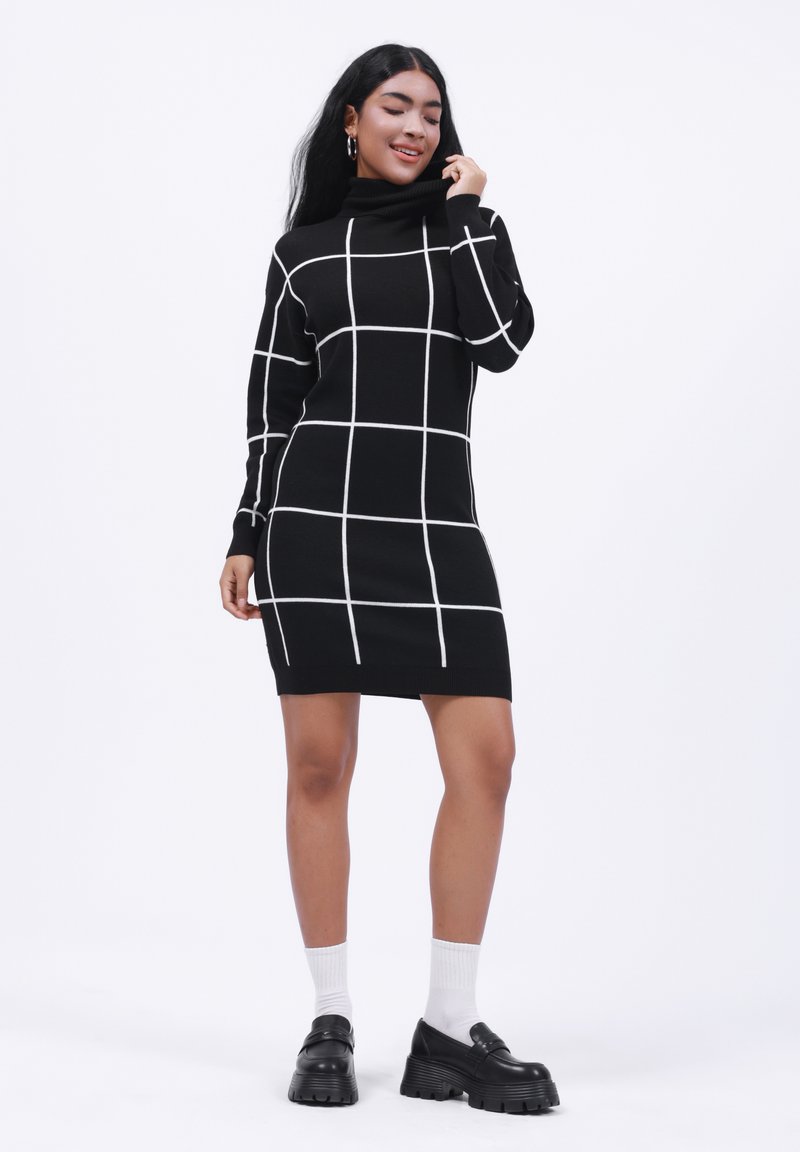 Melnas turtleneck kleitas ar baltu režģa rakstu, piegulošs dizains, ribota apkakle un izliektas maliņas. Modelis valkā masīvas melnas kurpes un baltas zeķes.
