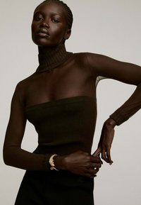 Top marrón ajustado con cuello alto acanalado, mangas largas transparentes y una textura suave. Presenta una delicada pulsera dorada en la muñeca.