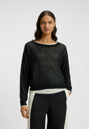Schwarzer, leichter, locker sitzender Pullover mit U-Boot-Ausschnitt, über einem weißen strukturierten Oberteil getragen. Lange Ärmel und subtile Transparenz.