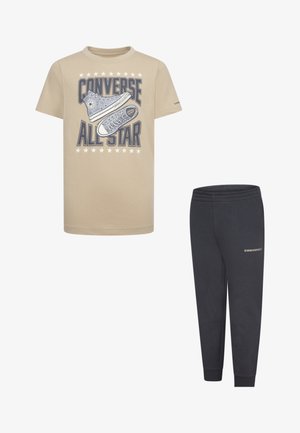 Beige T-Shirt mit einem blauen Converse All Star Grafik, kombiniert mit navyblauen Jogginghosen mit Logo. Beide haben eine lässige, lockere Passform.