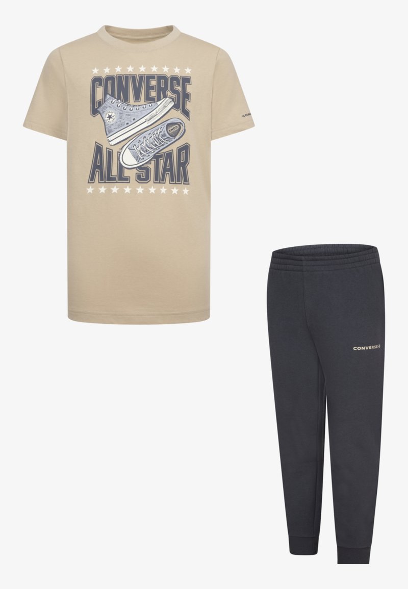 Beige T-shirt, kékkel nyomott Converse All Star grafikával, párosítva sötétkék melegítőnadrággal, amelyen logó található. Mindkettő laza, kényelmes szabású.