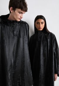 To personer har på seg løse, glinsende svarte ytterjakker med hette og snøring. Stoffet har en subtil tekstur og glans.