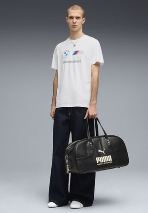 Młody mężczyzna w białej koszulce BMW Motorsport i ciemnych, szerokich dżinsach, trzymający czarną torbę sportową Puma na szarym tle.