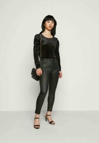 Camisa preta de manga longa com tecido texturizado e ombros volumosos, combinada com calças skinny pretas e sandálias de salto alto com tiras. Bolsa preta.