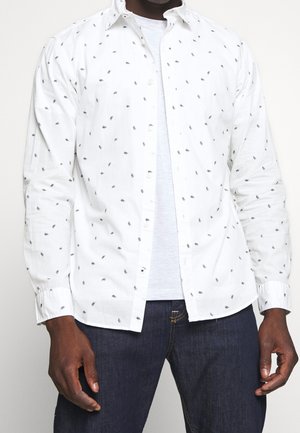 Mann trägt weißes Hemd mit kleinen schwarzen Blattmustern über weißem T-Shirt und dunkelblauer Jeans, steht vor einfarbigem Hintergrund.