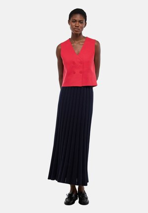 Femme portant un gilet rouge sans manches à double boutonnage, une longue jupe plissée bleu marine et des chaussures noires, se tenant devant un fond blanc uni.