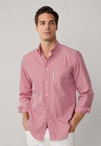 Homme avec des cheveux bruns portant une chemise à boutons rouge et blanche à rayures verticales, avec les manches retroussées, se tenant avec les mains dans les poches d'un pantalon blanc.