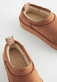 Barna slip-on mamusz, amely puha, texturált külső felülettel, plüss belső béléssel és szőtt címkészletével rendelkezik a sarkán. Gumi talp.
