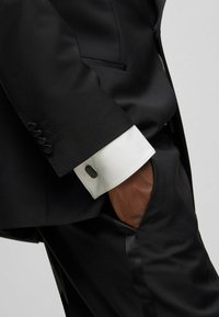 Completo nero con rever a punta, con un polsino bianco e un dettaglio con bottone nero. Tessuto dall'aspetto liscio con una vestibilità slim e design su misura.