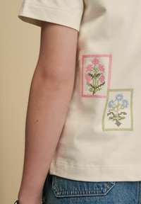 T-shirt crème à manches courtes avec deux écussons floraux rectangulaires brodés en rose et bleu, avec des détails verts sur un tissu texturé.