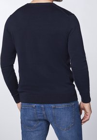 Marinblå långärmad tröja med rund halsringning, ribbade muddar och fåll. Bärs med ljusblå denimjeans och visar enkel design och textur.