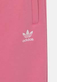 Růžová bavlněná mikina s výšivkou bílého loga Adidas, nad kterým jsou tři paralelní pruhy. Hladký pocit na dotek, s viditelnými detaily šití.