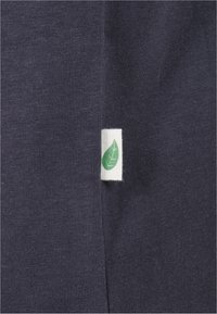 Tissu gris foncé avec une petite étiquette blanche comportant un symbole de feuille verte cousue dans une couture verticale.