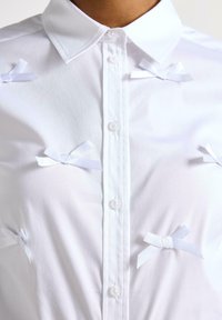 Camicia bianca con bottoni, caratterizzata da piccoli fiocchi di raso bianchi come dettagli, realizzata in un tessuto morbido con colletto classico e chiusura a bottoni.