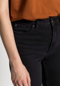 Jeans de mezclilla negra con una silueta ajustada, que cuentan con bolsillos frontales, un cierre de botón y una textura sutil. Combinados con una blusa marrón.