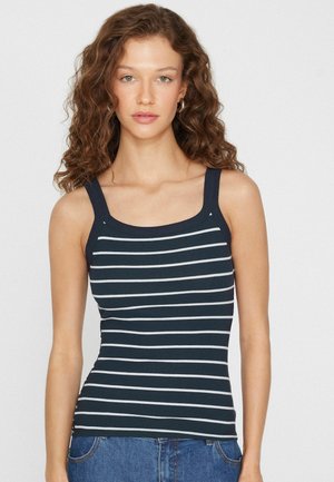 Junge Frau mit lockigem braunem Haar, trägt ein ärmelloses, marineblau-weiß gestreiftes Oberteil und blaue Jeans, schaut geradeaus vor einem einfarbigen Hintergrund.