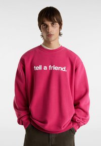 Růžová oversized mikina s bílým nápisem "řekni to kamarádovi." Kulatý výstřih, žebrované manžety, měkká textura, uvolněný střih, kombinováno s tmavými kalhotami.