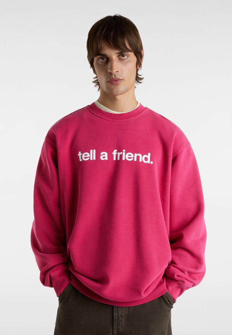 Lyserød oversized sweatshirt med hvid tekst "tell a friend." Rund hals, ribbede manchetter, blød tekstur, afslappet pasform, parret med mørke bukser.