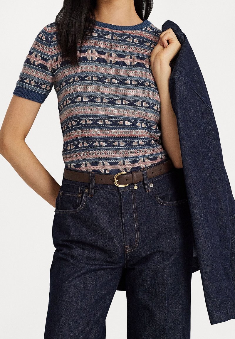 Femme portant un pull à manches courtes à motif, rentré dans un jean sombre taille haute avec une ceinture marron, tenant une veste en denim foncé sur une épaule.